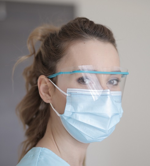 A photo of a light-skinned, feminine-presenting person with brown hair wearing medical eye protectors and a face mask.