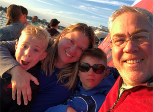 The Gramer Family on a boat, posing for a picture. From left to right: a young blonde boy, a middle-aged woman with brown hair, a young brown-haired boy with sunglasses, and a middle-aged man with gray hair and glasses.