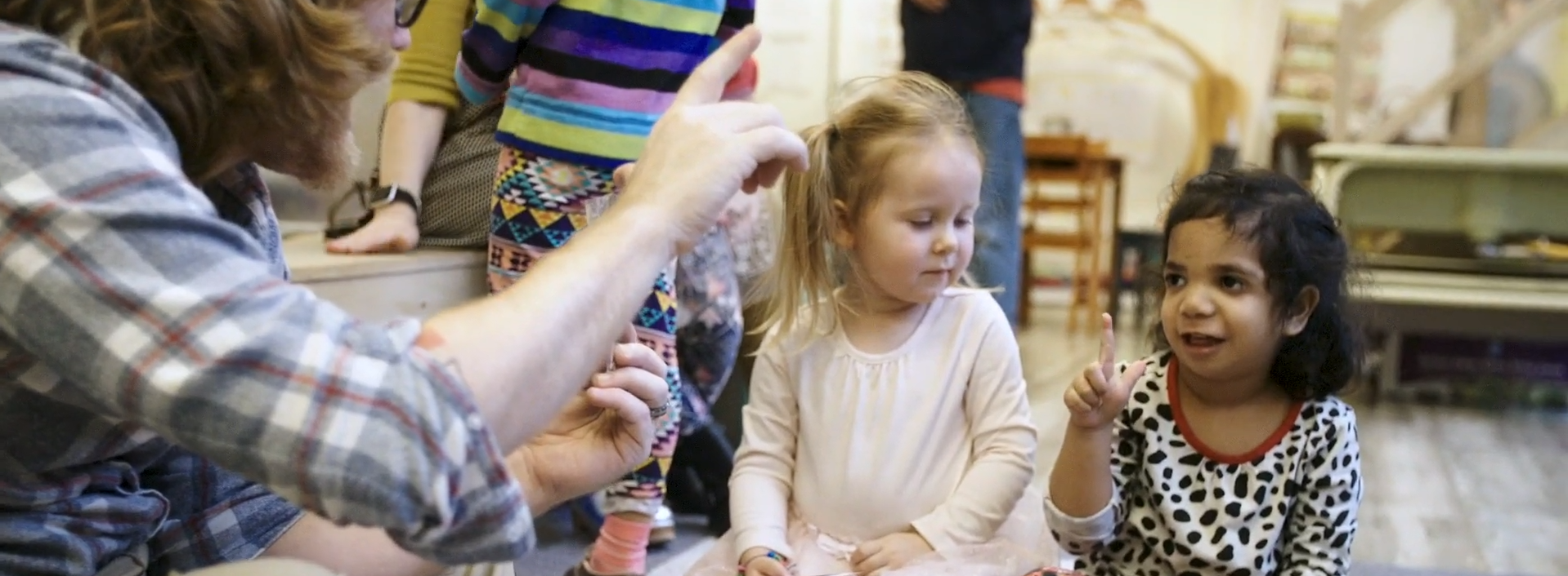 A photo of Rosen Family Preschool. In the foreground on the left, the arm of an instructor is signing to two sitting students on the right. The student on the left is looking at the student on the right, who is signing the letter L.