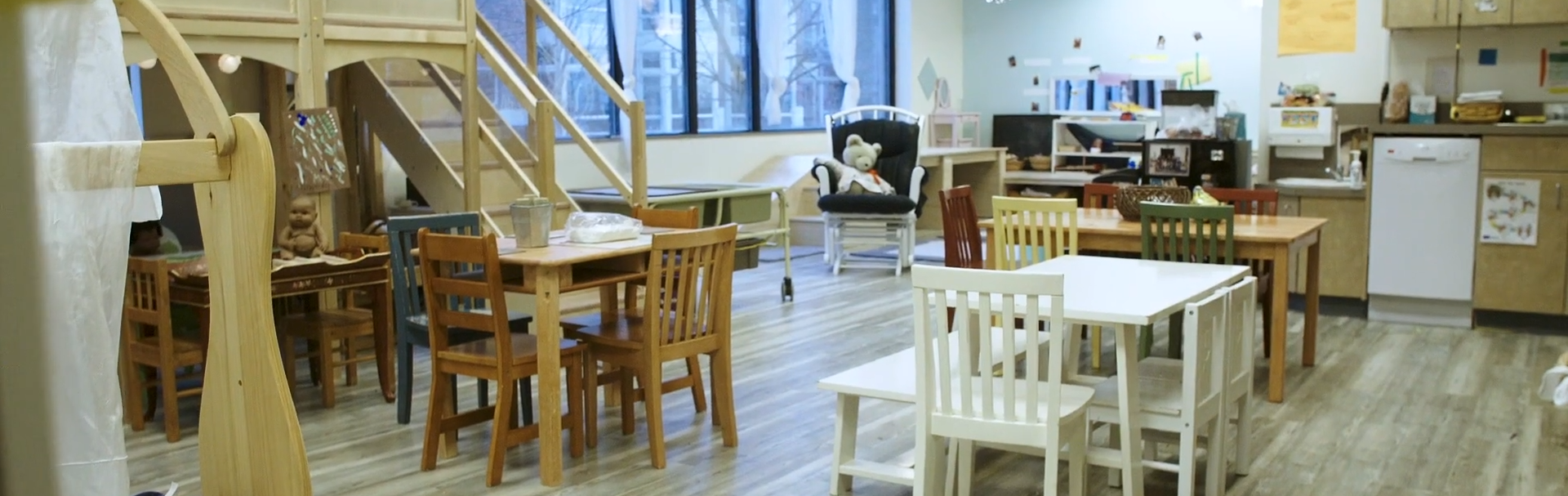 A photo of an empty Rosen Family Preschool. It is a bright classroom with a couple desks, loft, counter, cabinets, and friendly decorations. In the background is a long set of windows.