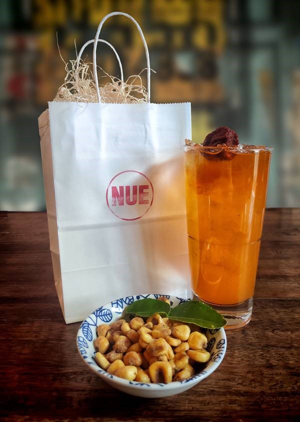 A photo of a bag, cocktail in a glass, and small bowl of nuts. The bag is paper, with the name Nue on the front. The orange cocktail is in a standard glass with ice. The nuts are in a blue and white bowl with a decorative leaf.