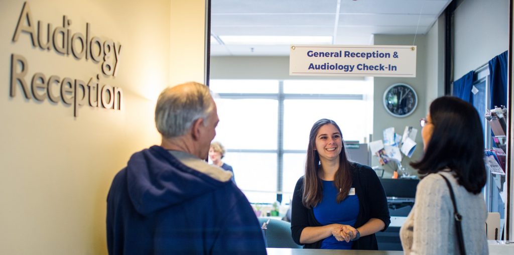 A photo of HSDC's front desk. The wall on the left says "Audiology Reception". Two people stand in front of the desk, looking at a smiling receptionist with long dark hair and a blue dress. Above, a sign says "General Reception & Audiology Check-In". Behind the desk is an office setting with a large window.